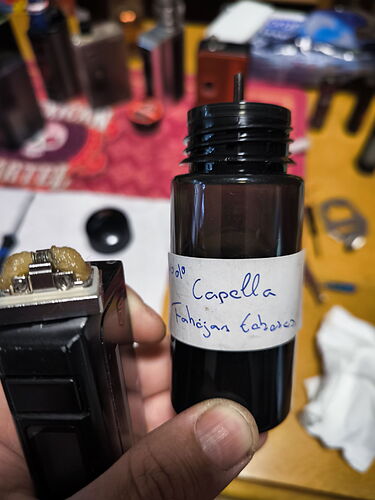 A hand holding a small vial labeled "Capella Michigan Sweet", with electronic cigarette or vaporizer parts scattered in the background. (Captioned with AI)