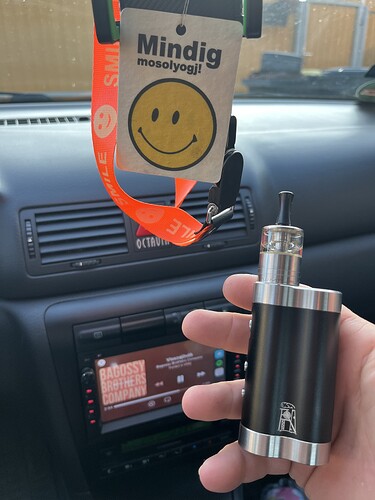Une main tenant une cigarette électronique noire à l'intérieur d'une voiture, derrière laquelle un emoji souriant est accroché avec un ruban orange sur lequel on peut lire « Souris toujours ! ». (Légendé par IA)
