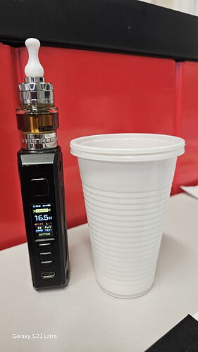 Une cigarette électronique noire et une tasse en plastique blanche sont visibles sur une table blanche, avec un fond rouge derrière. (Sous-titré par IA)