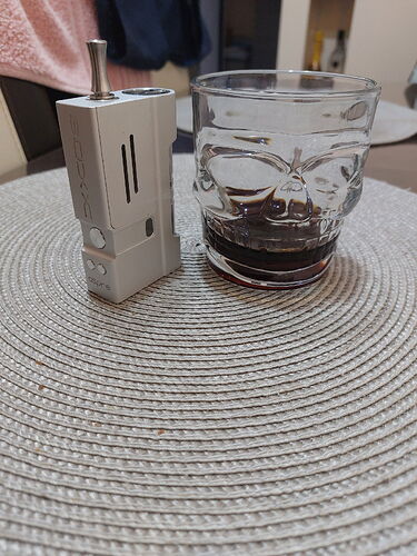 On the table, a grey vaporizer and a skull-patterned glass containing a dark drink are visible. (Captioned by AI)