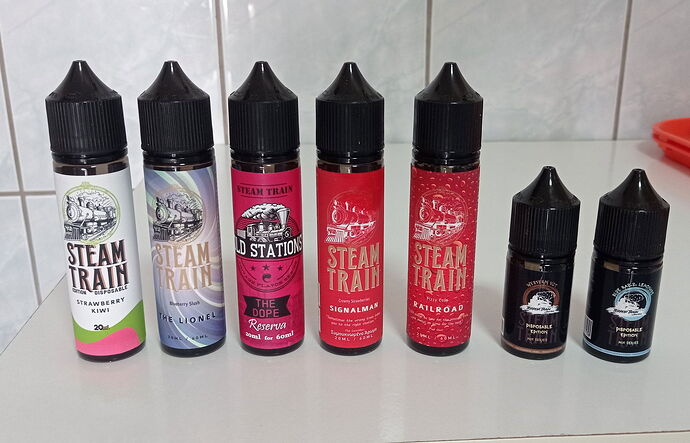 Az képen hét különböző Steam Train márkájú e-folyadék palack látható, különböző ízekkel és dizájnokkal egymás mellett, egy fehér asztalon. (Feliratozva AI-val)