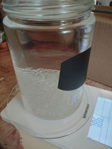 Egy átlátszó üveg befőttesüveg, benne szemcsés, áttetsző anyaggal, egy fehér digitális konyhai mérlegen áll, amelyen számok láthatók. (Feliratozva AI-val)