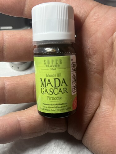 Une petite bouteille tenue dans une main, portant une étiquette « MADA GAS CAR » et la saveur « Pistachio ». (Légendé par IA)