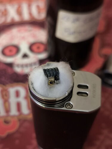 The image shows a top-down view of an electronic cigarette coil with cotton installed, and a partially blurred bottle with a label in the background. (Autocaptioned AI)