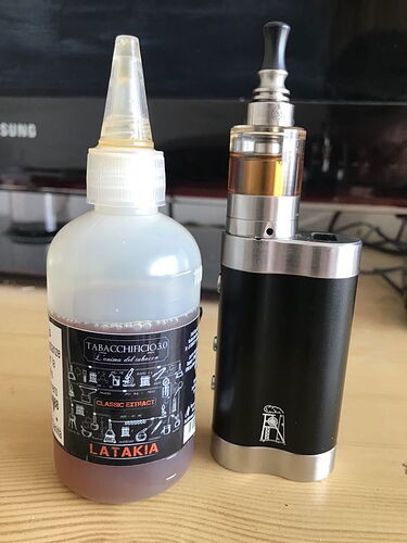A képen egy e-cigaretta mellett egy Tabacchificio 3.0 LATATIA dohányízesítő klasszikus kivonatával töltött üveg látható egy fa asztalon. (Feliratozva AI-val)