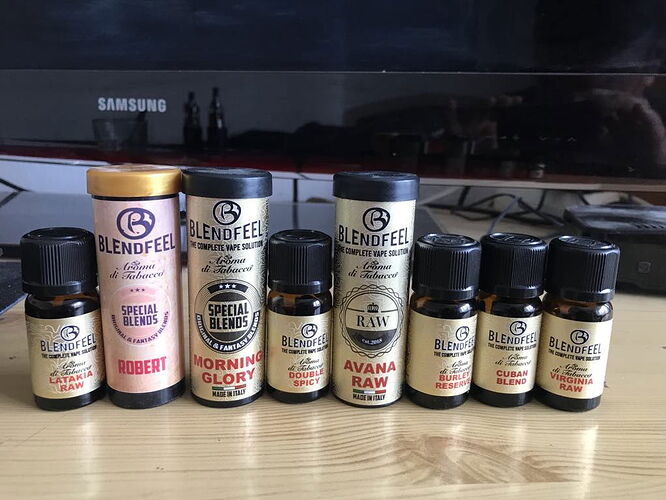 Az asztalon különböző Blendfeel márkájú, füstölőkhöz vagy vape folyadékokhoz tartozó aromaüvegek és dobozok sorakoznak egy Samsung monitor előtt. (Feliratozva AI-val)