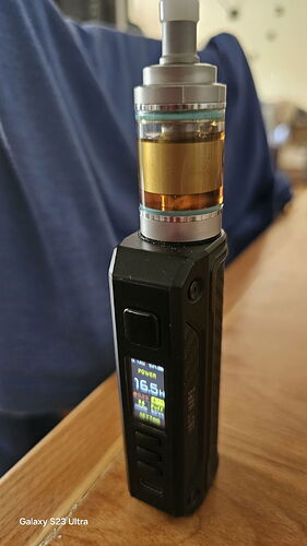 Sur la photo, un appareil cigarette électronique (vape) noir est visible sur une surface en bois, avec une personne vêtue de bleu en arrière-plan. (Légendé par IA)