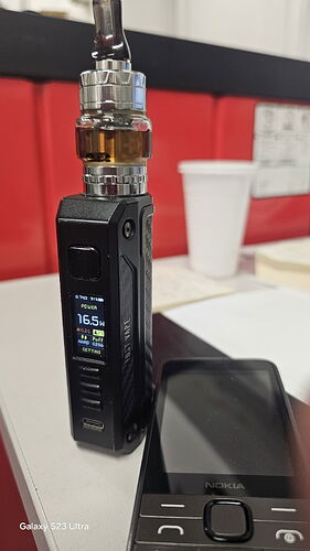 Une cigarette électronique noire et un vieux téléphone Nokia sont visibles sur la table, avec une tasse en papier blanche en arrière-plan. (Légendé par l'IA)