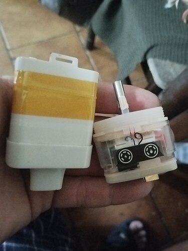 Two electronic components are held in a hand, one is yellow and white, and the other is transparent with internal parts. (Auto-captioned)