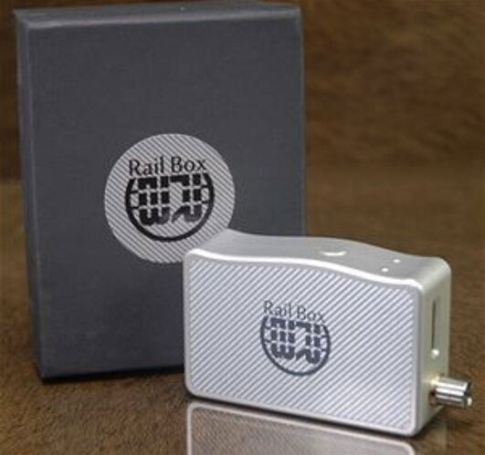 Egy ezüst színű, Rail Box feliratú elektronikus eszköz látható a sötétszürke csomagolódoboza mellett, melyen szintén a Rail Box logó szerepel. (Feliratozva AI-val)