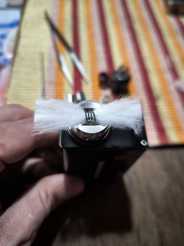 Egy kéz tart egy tekercselt huzalt kvarcot tartalmazó elektromos cigaretta (vape) porlasztó fejét, miközben a háttérben ecsetek és festékek láthatók egy csíkos felületen. (Feliratozva AI-val)