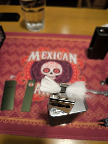 Az asztalon egy mexikói stílusú terítőn egy elektromos cigaretta, illetve különböző vaping eszközök láthatók, köztük egy fém vape és egy fehér vattának tűnő anyag. (Feliratozva AI-val)