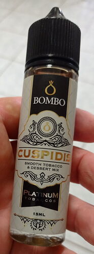 A képen egy 15 ml-es, Bombo márkájú, Cuspids nevű, sima dohány- és desszertkeverék ízű e-liquid palack látható. (Feliratozva AI-val)