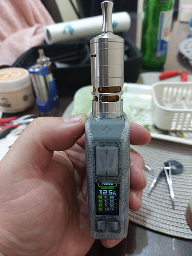 Une cigarette électronique (vape) tenue à la main, avec un écran numérique, est visible sur fond d'objets divers. (Légendé par l'IA)