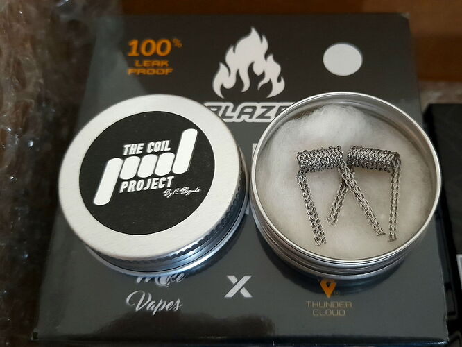 Az képen egy fémdoboz látható, amelyben két előre tekercselt fűtőtekercs helyezkedik el, mögötte pedig egy doboz áll, melyen több logó és a "100% LEAK PROOF" felirat olvasható. (Feliratozva AI-val)