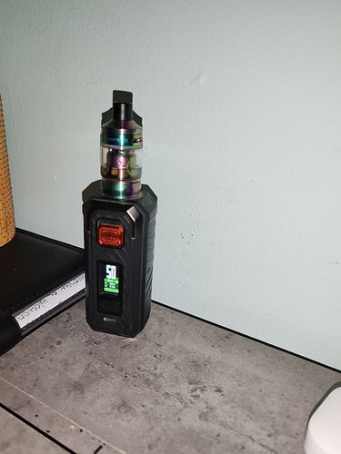 Une cigarette électronique noire avec un écran numérique (vape) est posée sur un coin de table devant un mur clair. (Sous-titré par IA)