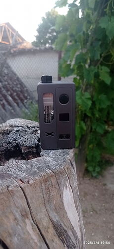 Egy fekete, kisméretű elektronikus eszköz helyezkedik el egy kidőlt, repedezett fatörzsön, háttérben zöld növények és egy kerítés látható. (Feliratozva AI-val)