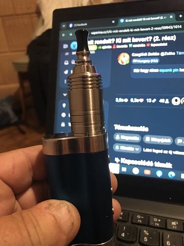 Egy személy egy kék és ezüst színű elektromos cigarettát tart a kezében egy laptop képernyője előtt. (Feliratozva AI-val)