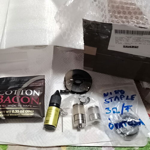 Divers accessoires de vapotage, tels que Cotton Bacon, des e-liquides et des pièces de rechange pour atomiseurs, sont disposés sur une surface à côté d'un colis. (Légendé par l'IA)