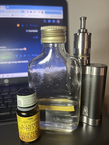 Sur une table, une bouteille à moitié pleine, une cigarette électronique et un flacon d'huile essentielle sont posés, avec l'écran d'un ordinateur portable en arrière-plan. (Sous-titré par IA)