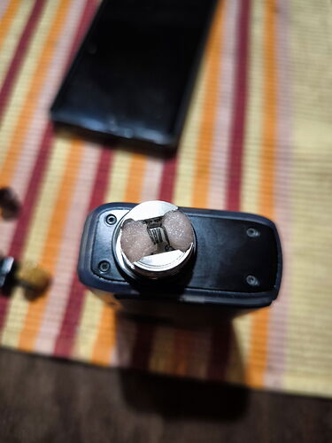 Egy elektromos cigaretta porlasztója egy fekete akkumulátoron, háttérben egy csíkos textília és egy fekete tok látható. (Feliratozva AI-val)