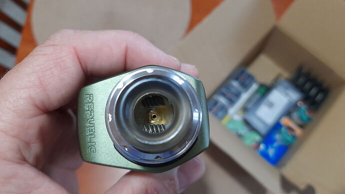 A hand holds a metal connector, showing contacts and coils inside, with boxes and batteries in the background. (Subtitled by AI)