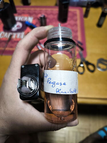 A hand holding a clear plastic bottle with a handwritten label, with various tools and instruments on a table behind it. (Caption provided by AI)