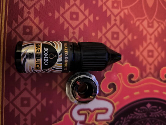 Une flasque de e-liquide saveur Glazed Donut est visible sur la photo, avec un bouchon métallique à côté, le tout devant un fond texturé. (Légendé par IA)