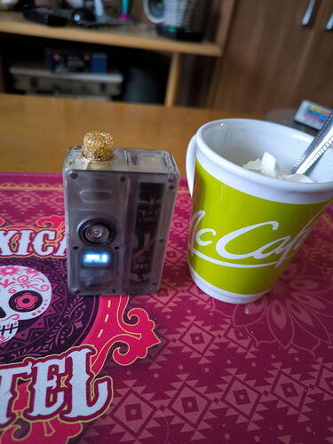 A képen egy átlátszó tokban lévő eszköz és egy zöld McCafé feliratos bögre látható, amely tejszínhabbal van töltve. (Feliratozva AI-val)