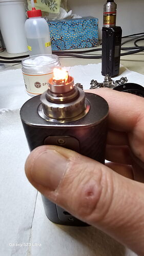 Une main tient un mod de vape allumé, avec divers accessoires et appareils de vapotage en arrière-plan. (Légendé par IA)