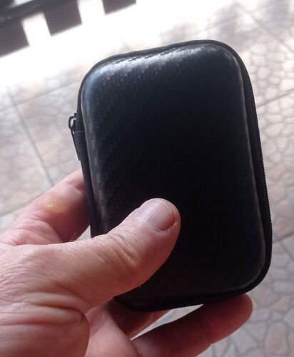 Egy kéz egy kis, fekete, cipzáras tokot tart, amelynek felülete szénszálas mintázatú. (Feliratozva AI-val)