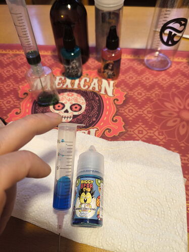 In the picture, a pipette full of blue liquid is held, next to which a colored bottle and various bottles are visible on a tablecloth. (Captioned by AI)