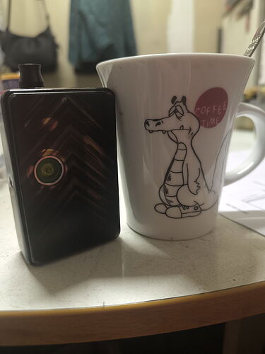 A képen egy fekete készülék és egy rajzos bögre látható, amelyen egy dínófigurával díszített címke található. (Feliratozva AI-val)