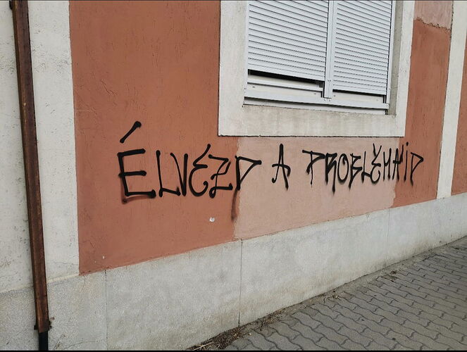 A sign painted in black paint on a brick-red wall reads "Enjoy your problems", with a window and a lowered shutter visible next to it. (Labeled by AI)