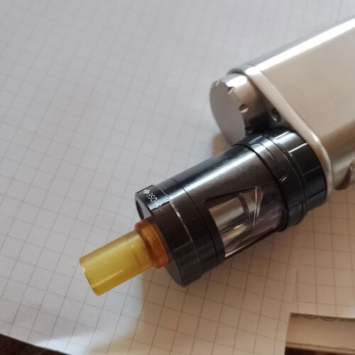 Une e-cigarette gris argenté avec un atomiseur sombre et un drip tip jaune translucide est visible sur un papier quadrillé. (Légendé par IA)