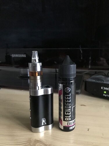 Az asztalon egy fekete ezüst elektronikais cigi és mellette egy Blendfeel Evening Cream nevű e-liquid áll egy televízió előtt. (Feliratozva AI-val)