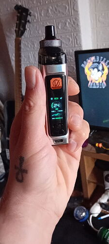 Une personne adulte tient un appareil de vapotage moderne dans sa main, avec divers objets et une télévision visibles en arrière-plan. (Légendé par IA)