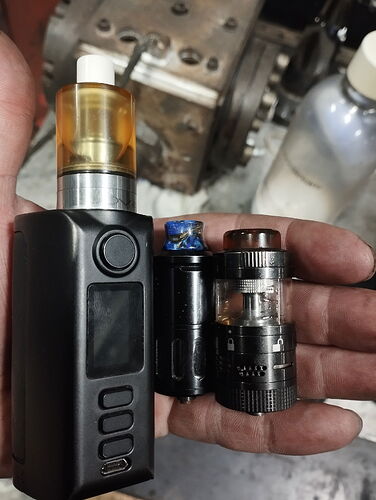 A képen egy kézben tartott vape eszköz látható, amely mellett különböző tartályok és alkatrészek is találhatók. (Feliratozva AI-val)