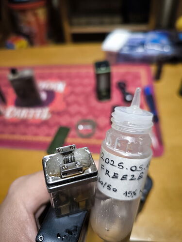 A hand holding an electronic cigarette part and a tank filled with liquid, with several other objects visible on a table in the background. (Caption provided by AI)